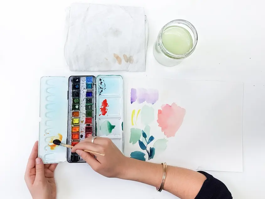 La palette aquarelle de Marie, un pinceau, un bocal et un chiffon pour réaliser les mélanges